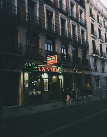 A dimly lit street scene in front of a café bar with a neon sign reading 'La Vera,' situated at the bottom of a building with classical architecture. People are walking on the sidewalk along the storefront.
