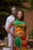 A couple stands together, with the woman in front wearing a vibrant traditional outfit featuring red, yellow, and green patterns. The man, dressed in a white garment with colorful accents, has his arms around her. The background has greenery and natural elements, creating a serene outdoor setting.