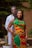 A couple stands together, with the woman in front wearing a vibrant traditional outfit featuring red, yellow, and green patterns. The man, dressed in a white garment with colorful accents, has his arms around her. The background has greenery and natural elements, creating a serene outdoor setting.