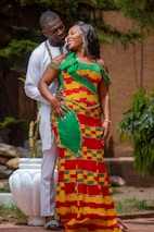 A couple is standing together outdoors, displaying affection and joy. The woman is wearing a colorful, traditional African dress with a vibrant pattern, while the man is dressed in a white outfit. They are surrounded by greenery, and a white pot is visible in the background.