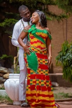 A couple is standing together outdoors, displaying affection and joy. The woman is wearing a colorful, traditional African dress with a vibrant pattern, while the man is dressed in a white outfit. They are surrounded by greenery, and a white pot is visible in the background.