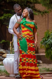 A couple is standing together outdoors, displaying affection and joy. The woman is wearing a colorful, traditional African dress with a vibrant pattern, while the man is dressed in a white outfit. They are surrounded by greenery, and a white pot is visible in the background.