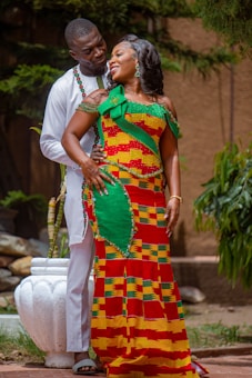 A couple is standing together outdoors, displaying affection and joy. The woman is wearing a colorful, traditional African dress with a vibrant pattern, while the man is dressed in a white outfit. They are surrounded by greenery, and a white pot is visible in the background.