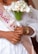 A close-up of a person wearing a white lace outfit and a sash that reads 'Bride' in glittery letters. They are holding a bouquet of white calla lilies tied with a ribbon. A diamond ring is visible on one hand, along with a gold wristwatch.