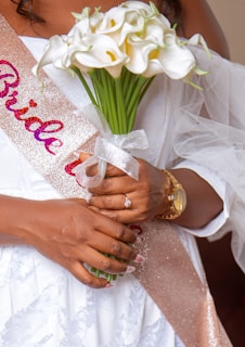 A close-up of a person wearing a white lace outfit and a sash that reads 'Bride' in glittery letters. They are holding a bouquet of white calla lilies tied with a ribbon. A diamond ring is visible on one hand, along with a gold wristwatch.