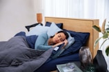 A person sleeps peacefully on a bed, surrounded by pillows and covered with a dark quilt. A wooden headboard and a side table with decorative items and a plant are visible. Soft, natural light enters through a sheer curtain.