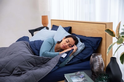 A person sleeps peacefully on a bed, surrounded by pillows and covered with a dark quilt. A wooden headboard and a side table with decorative items and a plant are visible. Soft, natural light enters through a sheer curtain.
