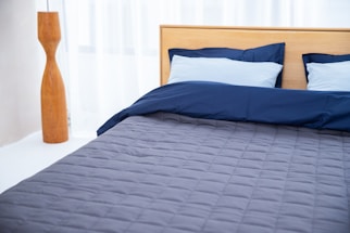 A neatly made bed with a wooden headboard is positioned against sheer white curtains. The bedding includes two light blue pillows and a navy blue duvet, with a gray quilted bedspread. To the left is a tall, wooden floor lamp with a sleek, modern design.