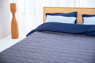 A neatly made bed with a wooden headboard is positioned against sheer white curtains. The bedding includes two light blue pillows and a navy blue duvet, with a gray quilted bedspread. To the left is a tall, wooden floor lamp with a sleek, modern design.