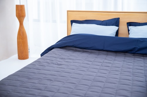 A neatly made bed with a wooden headboard is positioned against sheer white curtains. The bedding includes two light blue pillows and a navy blue duvet, with a gray quilted bedspread. To the left is a tall, wooden floor lamp with a sleek, modern design.