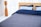A neatly made bed with a wooden headboard is positioned against sheer white curtains. The bedding includes two light blue pillows and a navy blue duvet, with a gray quilted bedspread. To the left is a tall, wooden floor lamp with a sleek, modern design.