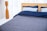 A neatly made bed with a wooden headboard is positioned against sheer white curtains. The bedding includes two light blue pillows and a navy blue duvet, with a gray quilted bedspread. To the left is a tall, wooden floor lamp with a sleek, modern design.
