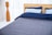 A neatly made bed with a wooden headboard is positioned against sheer white curtains. The bedding includes two light blue pillows and a navy blue duvet, with a gray quilted bedspread. To the left is a tall, wooden floor lamp with a sleek, modern design.