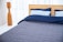 A neatly made bed with a wooden headboard is positioned against sheer white curtains. The bedding includes two light blue pillows and a navy blue duvet, with a gray quilted bedspread. To the left is a tall, wooden floor lamp with a sleek, modern design.