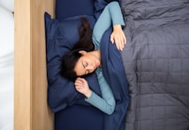 A person is peacefully sleeping in a bed, covered with a dark blue blanket. The bed is positioned near a wooden headboard, and the person is lying on a matching blue pillow. They are wearing a light blue long-sleeve shirt, with one arm resting on their pillow and the other under the blanket.