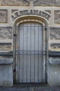 An old stone doorway featuring wrought iron bars across the entrance. The structure above the door has the name 'A.R. Delamater' engraved in large, ornate letters. The stone has a weathered appearance, indicating age, and the ground in front is paved.