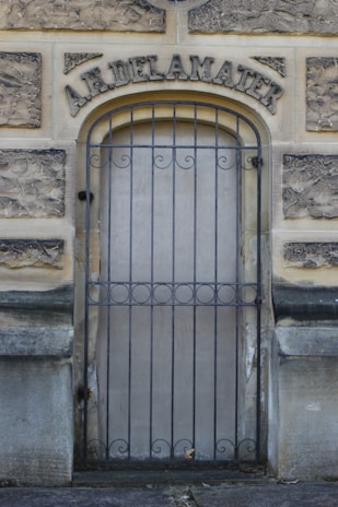 An old stone doorway featuring wrought iron bars across the entrance. The structure above the door has the name 'A.R. Delamater' engraved in large, ornate letters. The stone has a weathered appearance, indicating age, and the ground in front is paved.