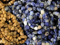 A close-up of various polished stones strung together in necklaces, featuring an assortment of blue and brown tones. The stones have a glossy finish, highlighting their smooth, rounded shapes.