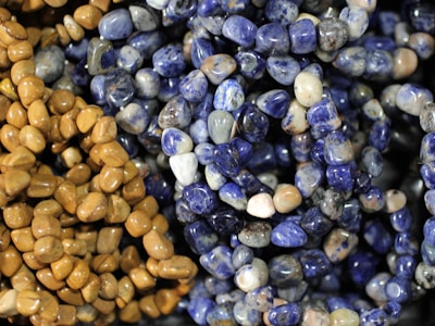 A close-up of various polished stones strung together in necklaces, featuring an assortment of blue and brown tones. The stones have a glossy finish, highlighting their smooth, rounded shapes.