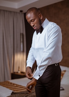 A man wearing a formal white shirt and black trousers is adjusting his cufflinks in a room. The room has a neatly made bed with patterned sheets and drapes partially covering a window.