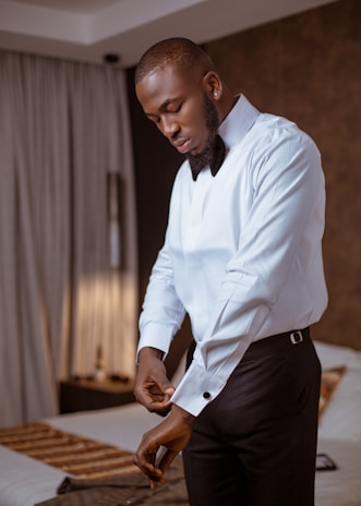 A man in a crisp white shirt and black trousers adjusting his watch in a minimalist room.