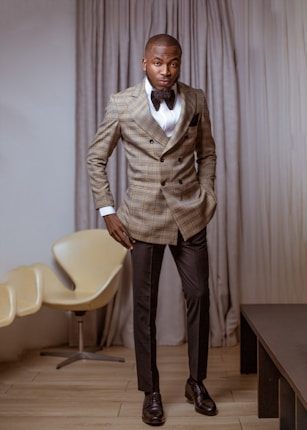 A man is standing in a well-furnished room, dressed in a checked double-breasted blazer, white dress shirt, black pants, and a bow tie. He has one hand in his pocket and is looking confidently towards the camera. Behind him, there is a modern, beige armchair and a curtain covering the backdrop.