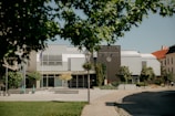 A modern university building surrounded by trees and a paved pathway. The architecture includes sleek lines with a combination of gray and white panels. Scooters are parked near the entrance, suggesting a mix of technology and transportation. The foreground features a grassy lawn with trees partially obscuring the view.