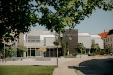 A modern university building surrounded by trees and a paved pathway. The architecture includes sleek lines with a combination of gray and white panels. Scooters are parked near the entrance, suggesting a mix of technology and transportation. The foreground features a grassy lawn with trees partially obscuring the view.