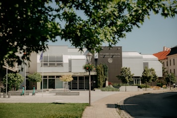 A modern university building surrounded by trees and a paved pathway. The architecture includes sleek lines with a combination of gray and white panels. Scooters are parked near the entrance, suggesting a mix of technology and transportation. The foreground features a grassy lawn with trees partially obscuring the view.
