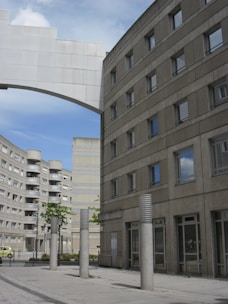 The image features an urban scene with modern, geometric buildings constructed from concrete. The architecture is characterized by clean lines and numerous windows. Tall cylindrical pillars line the walkway in the foreground, and a white arch connects two of the buildings. The sky is partly cloudy, adding depth to the scene.