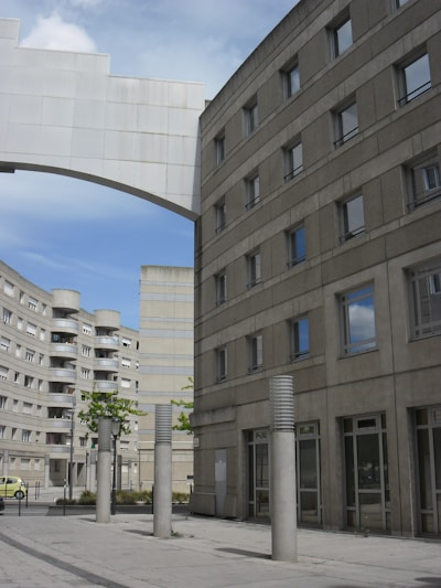 The image features an urban scene with modern, geometric buildings constructed from concrete. The architecture is characterized by clean lines and numerous windows. Tall cylindrical pillars line the walkway in the foreground, and a white arch connects two of the buildings. The sky is partly cloudy, adding depth to the scene.