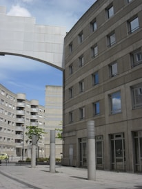 The image features an urban scene with modern, geometric buildings constructed from concrete. The architecture is characterized by clean lines and numerous windows. Tall cylindrical pillars line the walkway in the foreground, and a white arch connects two of the buildings. The sky is partly cloudy, adding depth to the scene.