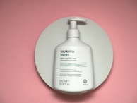 A white plastic pump bottle, labeled with the brand name 'Sesderma Salises' and described as a foamy soap-free cream. The bottle is positioned on a circular white platform against a pink background.