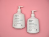 Two white bottles of Sesderma Salises foamy soap-free cream with pump dispensers are placed on a pastel pink background. The bottles are labeled in English and Spanish, indicating they are for acne-prone skin cleansing and contain salicylic acid.