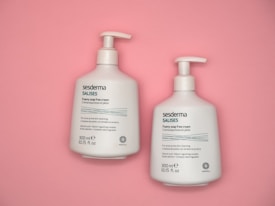 Two white bottles of Sesderma Salises foamy soap-free cream with pump dispensers are placed on a pastel pink background. The bottles are labeled in English and Spanish, indicating they are for acne-prone skin cleansing and contain salicylic acid.