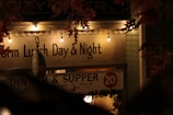 A vintage storefront sign illuminated by warm, ambient string lights. The sign advertises lunch and dinner services with a visible pricing of 20 cents. Autumn leaves frame the top of the image, enhancing the nostalgic atmosphere. An old-fashioned clock hangs prominently in the foreground.