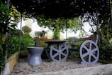 A rustic wooden bench with wheel-like supports is positioned in a garden setting. The bench and a matching stool rest on gravel, surrounded by lush green foliage. Vines create a natural canopy overhead, casting shadows on the seating area. The background reveals more greenery and hints of a bright sky.