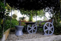 A rustic wooden bench with wheel-like supports is positioned in a garden setting. The bench and a matching stool rest on gravel, surrounded by lush green foliage. Vines create a natural canopy overhead, casting shadows on the seating area. The background reveals more greenery and hints of a bright sky.