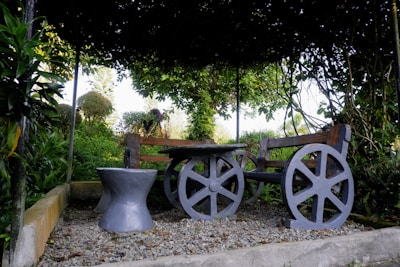 A rustic wooden bench with wheel-like supports is positioned in a garden setting. The bench and a matching stool rest on gravel, surrounded by lush green foliage. Vines create a natural canopy overhead, casting shadows on the seating area. The background reveals more greenery and hints of a bright sky.