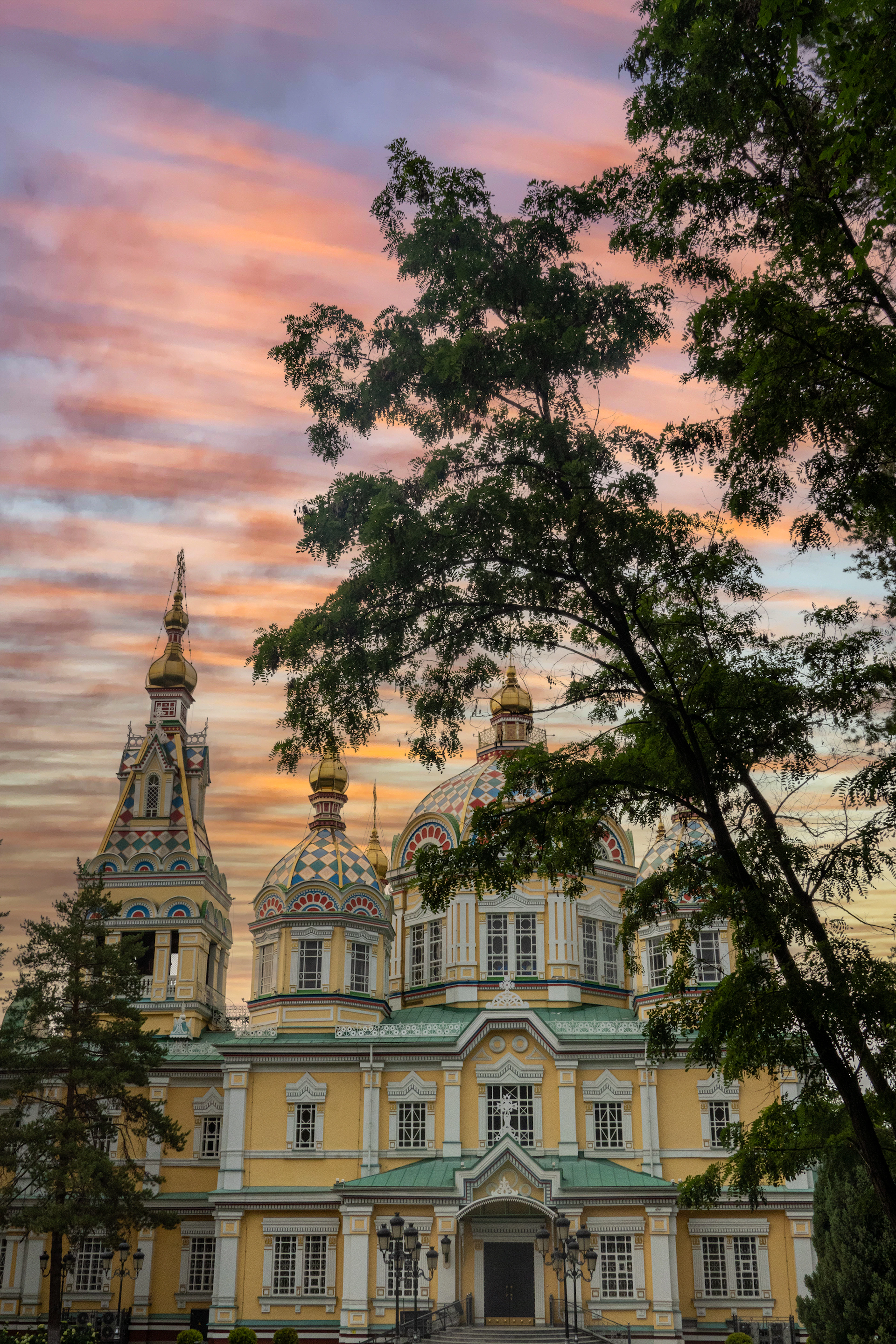 Ascension Cathedral's golden domes rise through tree branches against a dramatic pink-and-orange sunset sky.
