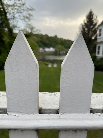 A close-up of a white picket fence with two pointed tops fills the foreground. In the background, a well-kept lawn leads to a waterbody surrounded by trees and distant houses under a cloudy sky. The scene gives a sense of a peaceful suburban setting.