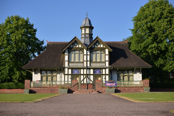 A quaint, traditional building with a dark, steeply pitched roof and timber-framed architecture sits at the end of a pathway. The facade features a symmetrical design with a central doorway flanked by windows and a small balcony above. Two large trees frame the building on either side, enhancing the picturesque setting. A banner is displayed on the railing, adding a touch of modernity to the historical aesthetics.