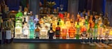A well-stocked bar counter features a diverse array of liquor bottles, with bright lighting highlighting the assortment. Various brands of whiskey, gin, and other spirits are displayed. Row of wine bottles is seen on the left side, while several upside-down wine glasses are neatly arranged on the right.
