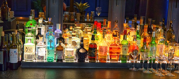A well-stocked bar counter features a diverse array of liquor bottles, with bright lighting highlighting the assortment. Various brands of whiskey, gin, and other spirits are displayed. Row of wine bottles is seen on the left side, while several upside-down wine glasses are neatly arranged on the right.