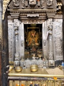 A richly decorated temple interior with an intricately carved doorway framing a deity statue adorned with colorful flower garlands. The scene includes offerings such as brass vessels and oil lamps on a wooden platform at the entrance.