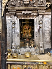 A richly decorated temple interior with an intricately carved doorway framing a deity statue adorned with colorful flower garlands. The scene includes offerings such as brass vessels and oil lamps on a wooden platform at the entrance.