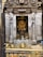 A richly decorated temple interior with an intricately carved doorway framing a deity statue adorned with colorful flower garlands. The scene includes offerings such as brass vessels and oil lamps on a wooden platform at the entrance.