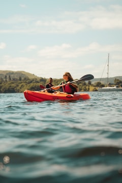 A person is kayaking on a calm body of water with hills and trees in the background. Another person is visible in the distance, also paddling. A sailboat is anchored nearby, and the sky is partly cloudy.