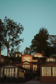 A series of modern, wooden cabins are nestled on a wooded hillside under a clear blue sky. Tall trees frame the scene, adding a natural backdrop to the structures, which feature large windows and glass panels. The warm sunlight casts gentle shadows, enhancing the serene atmosphere.