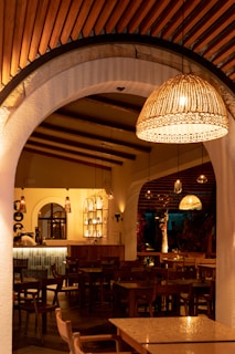 A cozy interior of a restaurant featuring wooden tables and chairs arranged neatly. Warm, ambient lighting is provided by ornate pendant lamps hanging from the ceiling. The arched entryways and ceiling beams enhance the elegant aesthetic, with decor that combines rustic and contemporary elements.